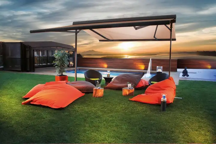 Abri de terrasse sur pied central au-dessus de poufs orange, près d’une piscine, au coucher du soleil