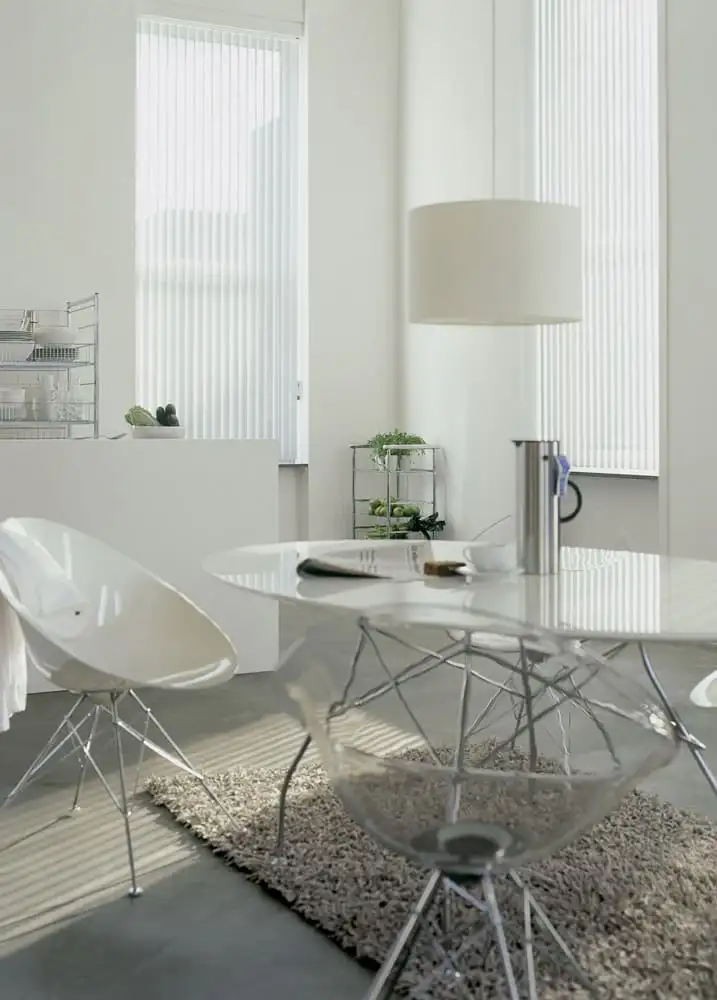 Stores à bandes verticales blancs devant les baies vitrées d’une cuisine claire, une table ronde et des chaises transparentes au premier plan