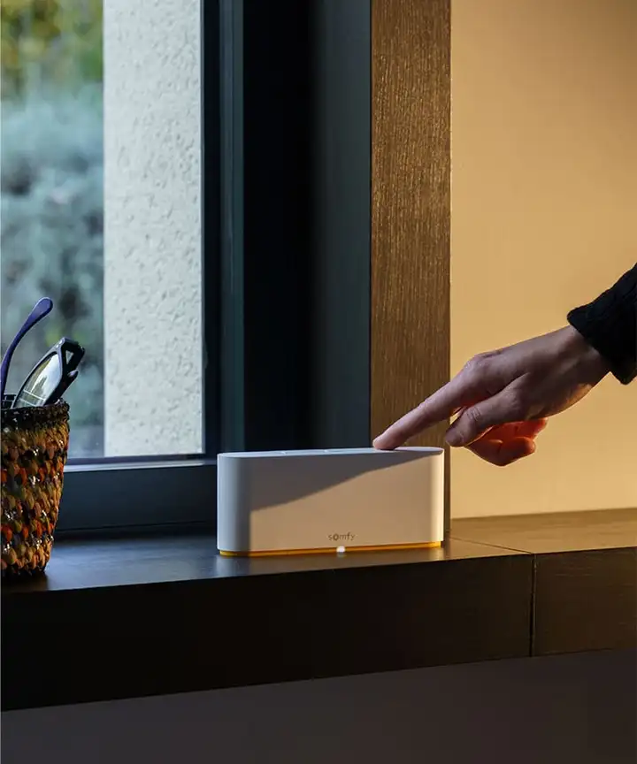 Main qui appuie sur une box domotique posée sur un meuble, près d’une fenêtre