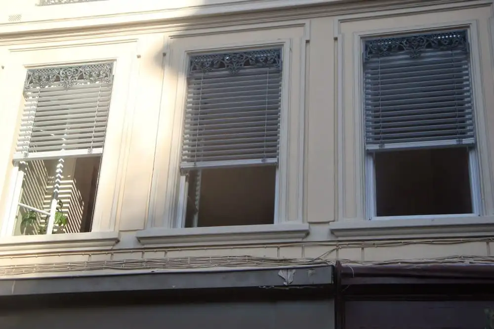 Façade d’immeuble lyonnais : lames orientables gris clair baissées derrière les cantonnières en fonte de trois fenêtres