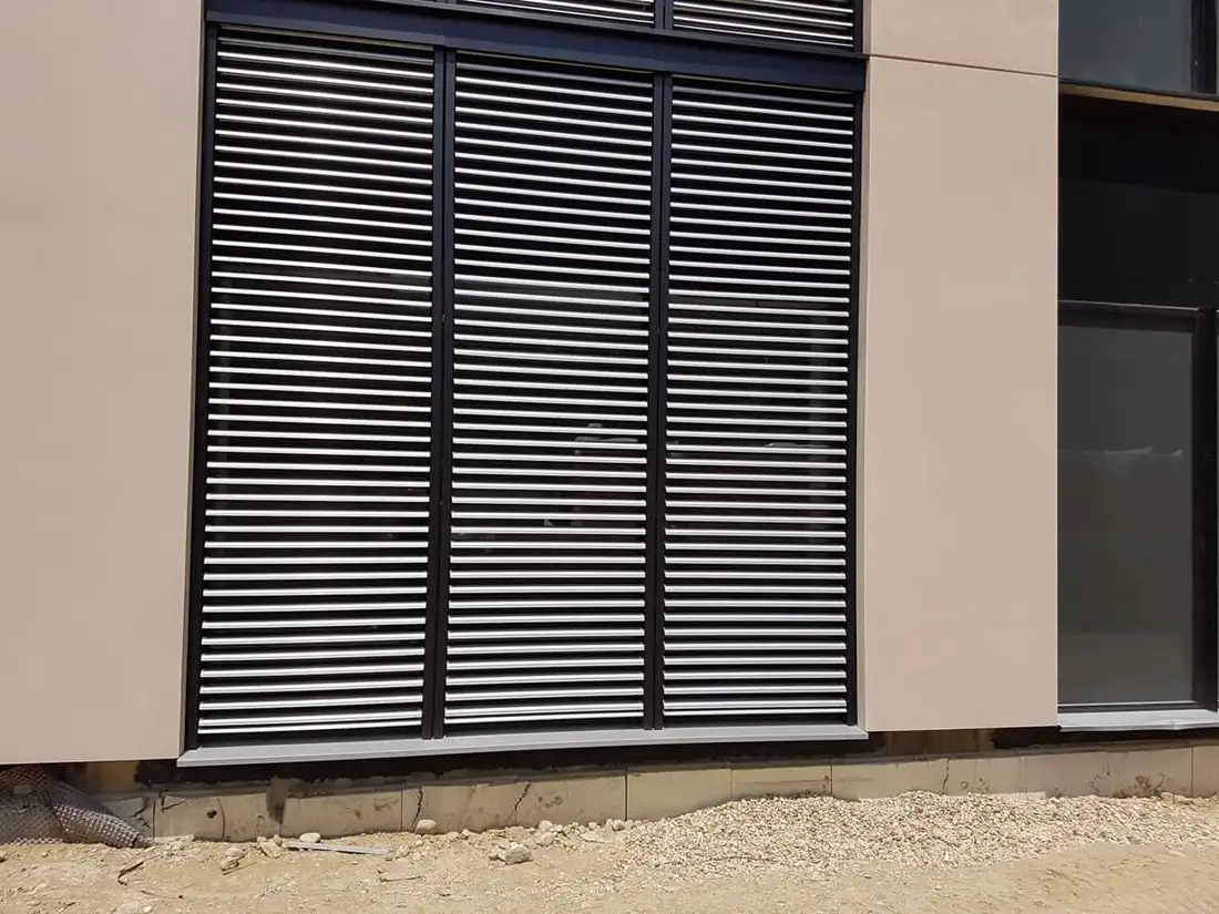 Brise-soleil orientables baissés devant une baie, lames d’aluminium entrouvertes, sur une façade claire