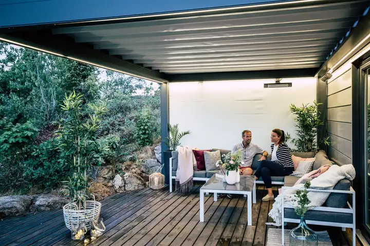 Pergola à toile adossée au bardage d’une maison, fermée au fond par un screen blanc, un couple assis dessous