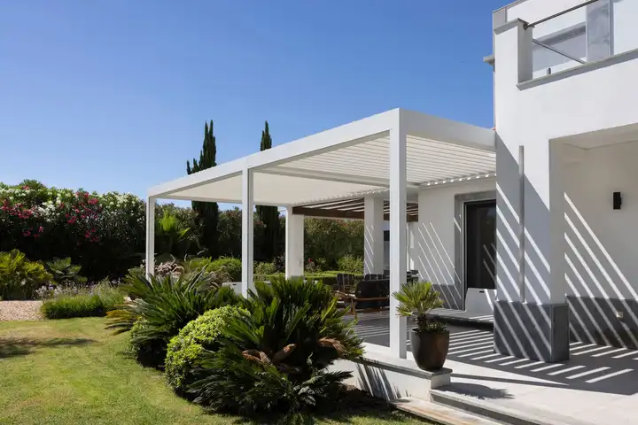 Pergola blanche à lames adossée à une maison blanche, son ombre sur la terrasse, dans un jardin méditerranéen