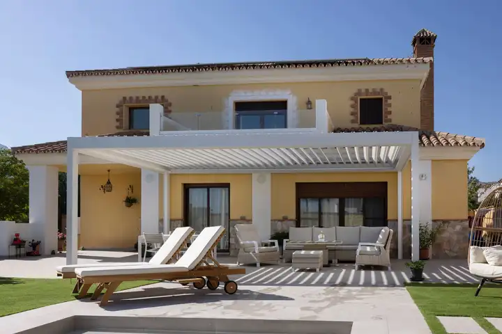 Pergola blanche à lames adossée à une villa ocre, transats et salon d’extérieur dessous