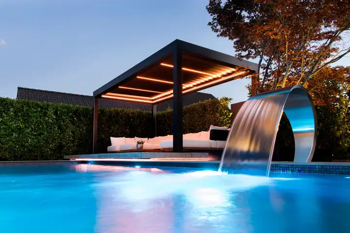 Pergola à lames éclairée le soir par des bandes lumineuses, au bord d’une piscine