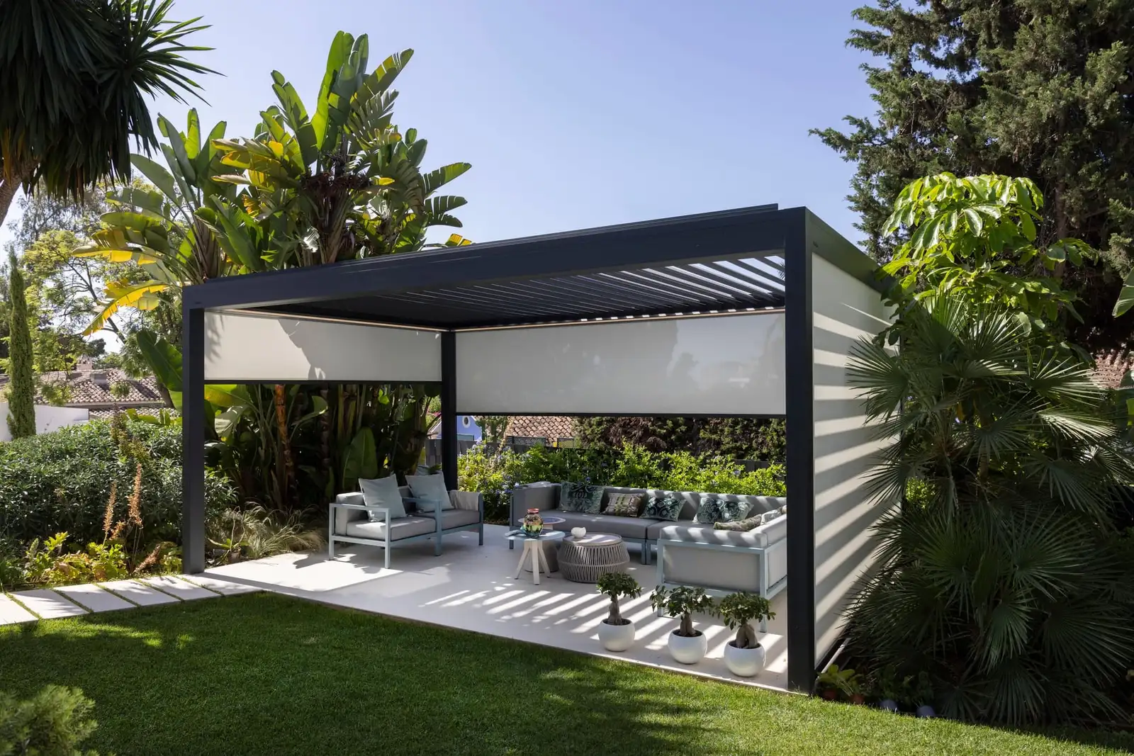 Pergola anthracite à lames posée sur une pelouse, un salon d’extérieur dessous, entre les palmiers d’un jardin