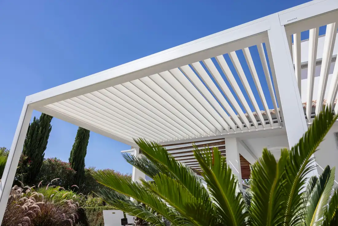 Toit d’une pergola blanche vu de dessous, ses lames ouvertes sur le ciel bleu