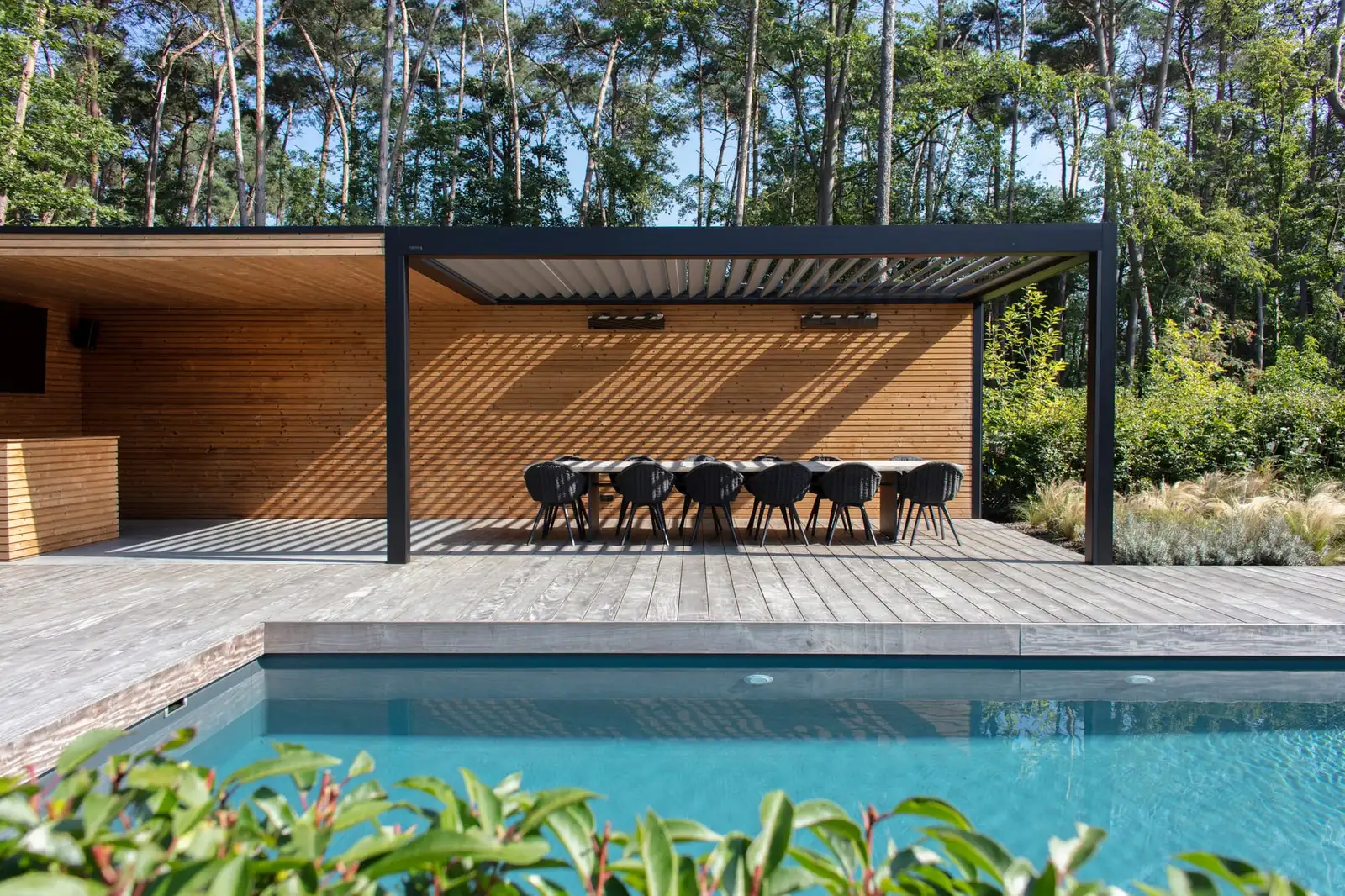 Pergola bioclimatique anthracite au bord d’une piscine, une longue table dressée sous son toit de lames