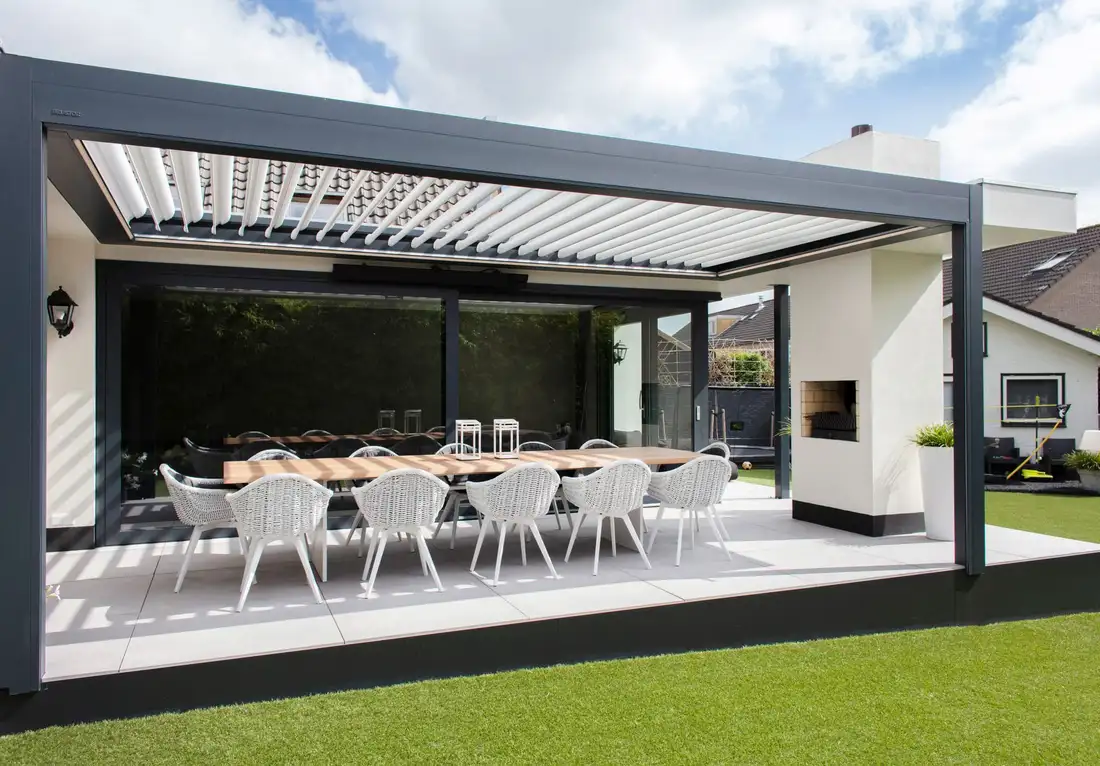 Pergola à lames blanches sur une terrasse de jardin, une longue table dressée dessous, près d’une cheminée d’extérieur