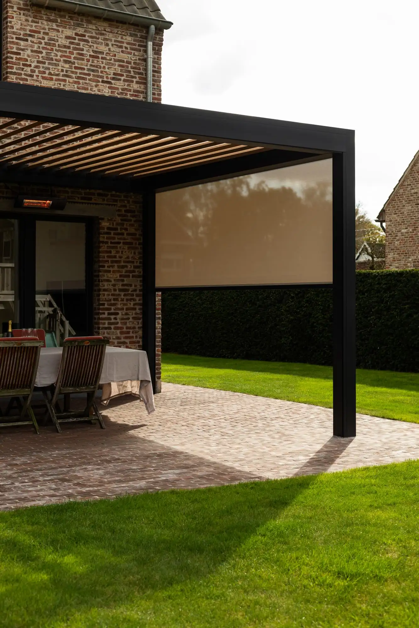 Pergola à lames anthracite adossée à une maison en briques, un screen déroulé au-dessus de la table