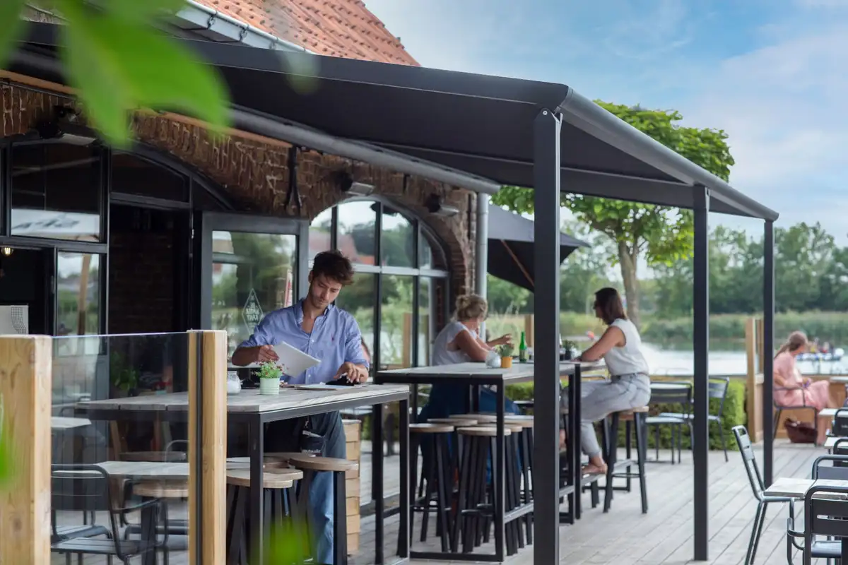 Terrasse de café au bord de l’eau sous une pergola à toile anthracite, clients attablés