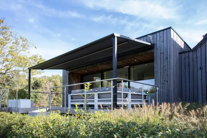 Pergola à toile anthracite au-dessus de la terrasse d’une maison en bois, guirlande lumineuse sous la toile