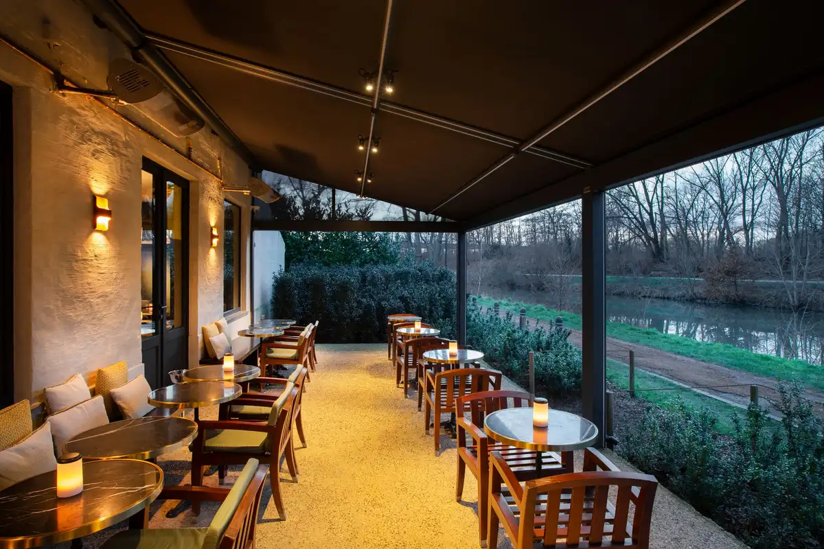 Terrasse de restaurant couverte par une pergola à toile adossée à un bâtiment en pierre, tables dressées le long d’un canal