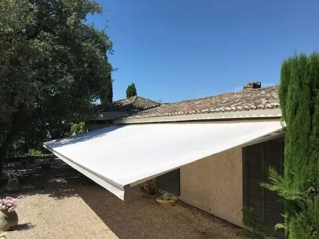 Store banne à toile blanche neuve, déployé au-dessus d’une terrasse bordée de cyprès, à Chaponost