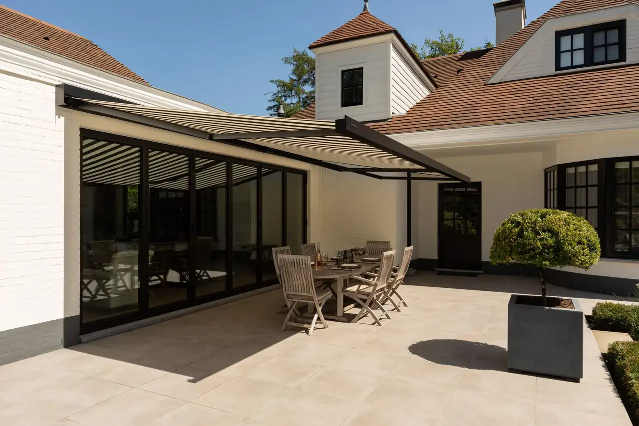 Store banne coffre à toile rayée grise déployé devant les baies d’une maison, au-dessus d’une terrasse dallée