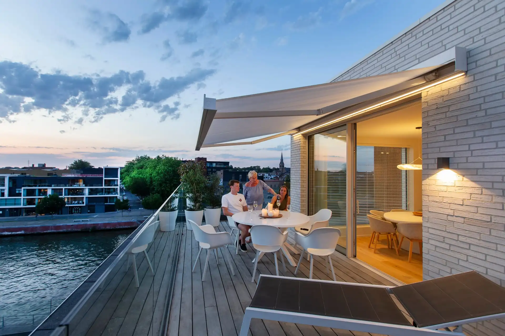 Terrasse d’appartement au crépuscule, au-dessus de l’eau, abritée par un store banne blanc éclairé par des LED