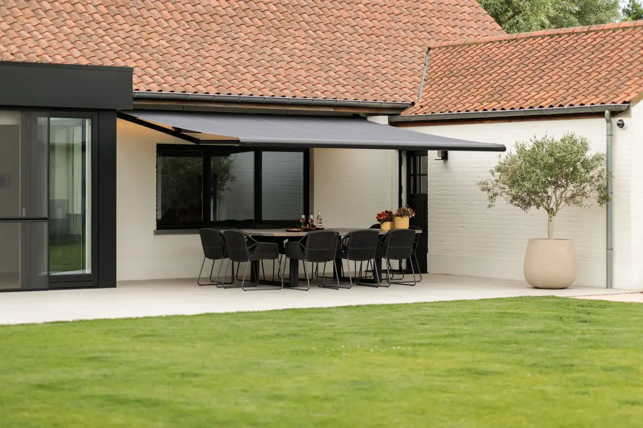 Store banne gris déployé au-dessus d’une grande table de jardin, devant une maison au bord d’une pelouse