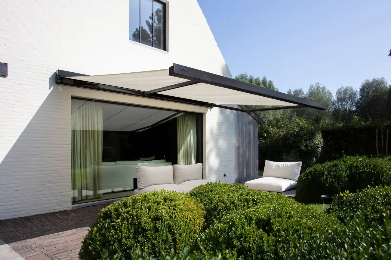 Store banne à toile claire déployé devant la grande baie vitrée d’une maison blanche, au-dessus d’un salon de jardin