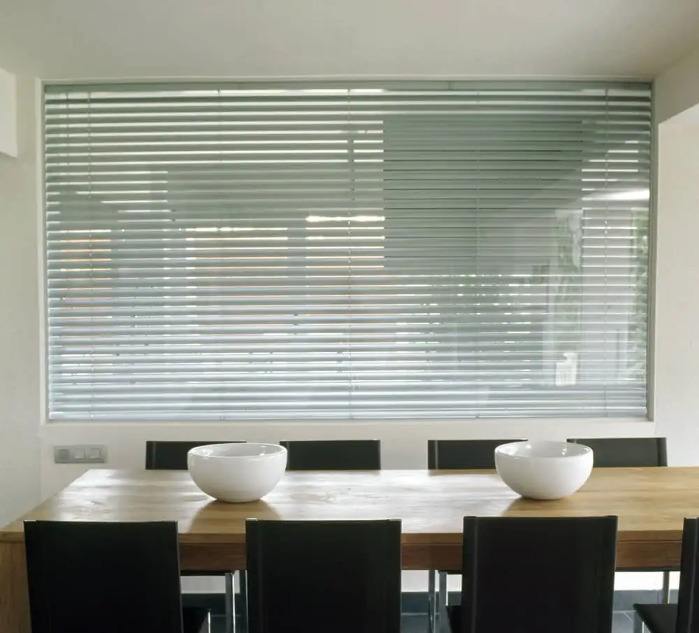 Store vénitien à lames d’aluminium clair baissé devant une large fenêtre, au-dessus d’une table en bois