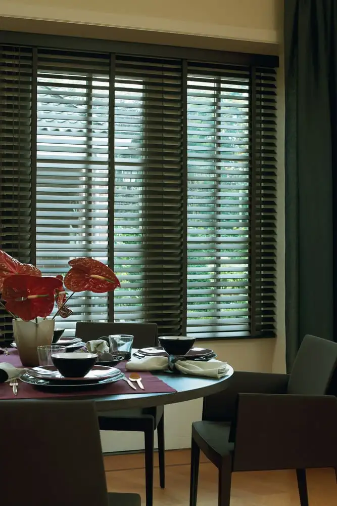 Store vénitien à lames de bois sombre devant la fenêtre d’une salle à manger, des anthuriums rouges sur la table
