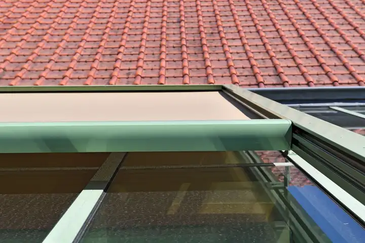 Coffre vert d’un store extérieur fixé en haut de la toiture vitrée d’une véranda, sous le rebord du toit