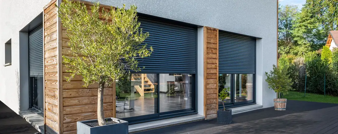 Maison contemporaine aux volets roulants gris anthracite, baissés à mi-hauteur derrière un olivier